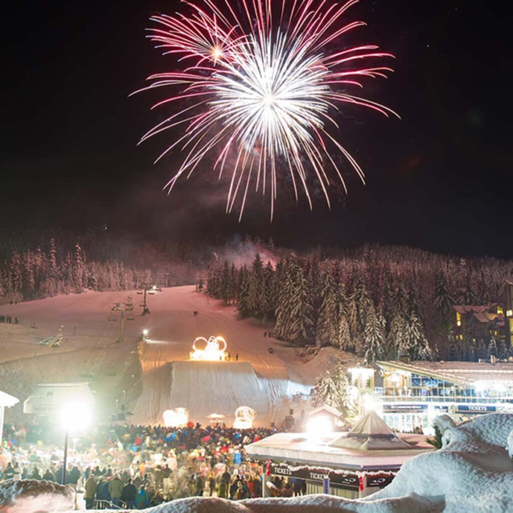 A large crowd of people watches fireworks above a ski jump with flaming rings. 